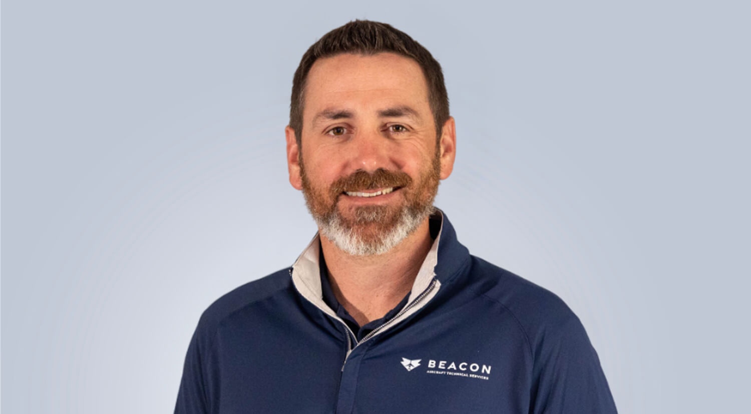 Barbour Updated Photo (1) A man with short brown hair and a beard, wearing a navy blue pullover with a BEACON logo, stands smiling against a light gray background.