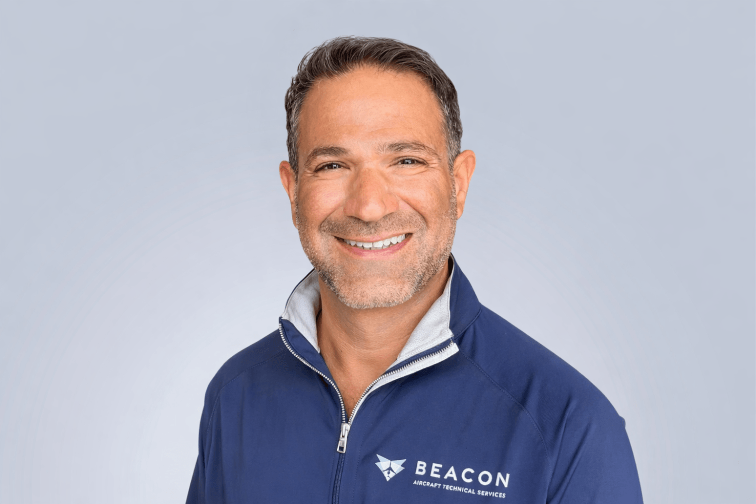A man with short brown hair and a trimmed beard smiles at the camera. He wears a navy blue zip-up jacket with BEACON Aircraft Technical Services and a paper plane logo on the chest, against a light gray background.
