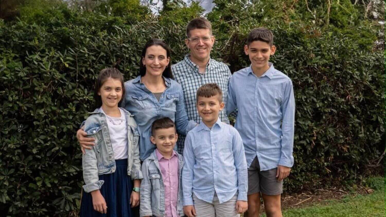 A family of six, including two adults and four children, stands outside in front of green bushes. They are all smiling and dressed in casual, light-colored clothing.