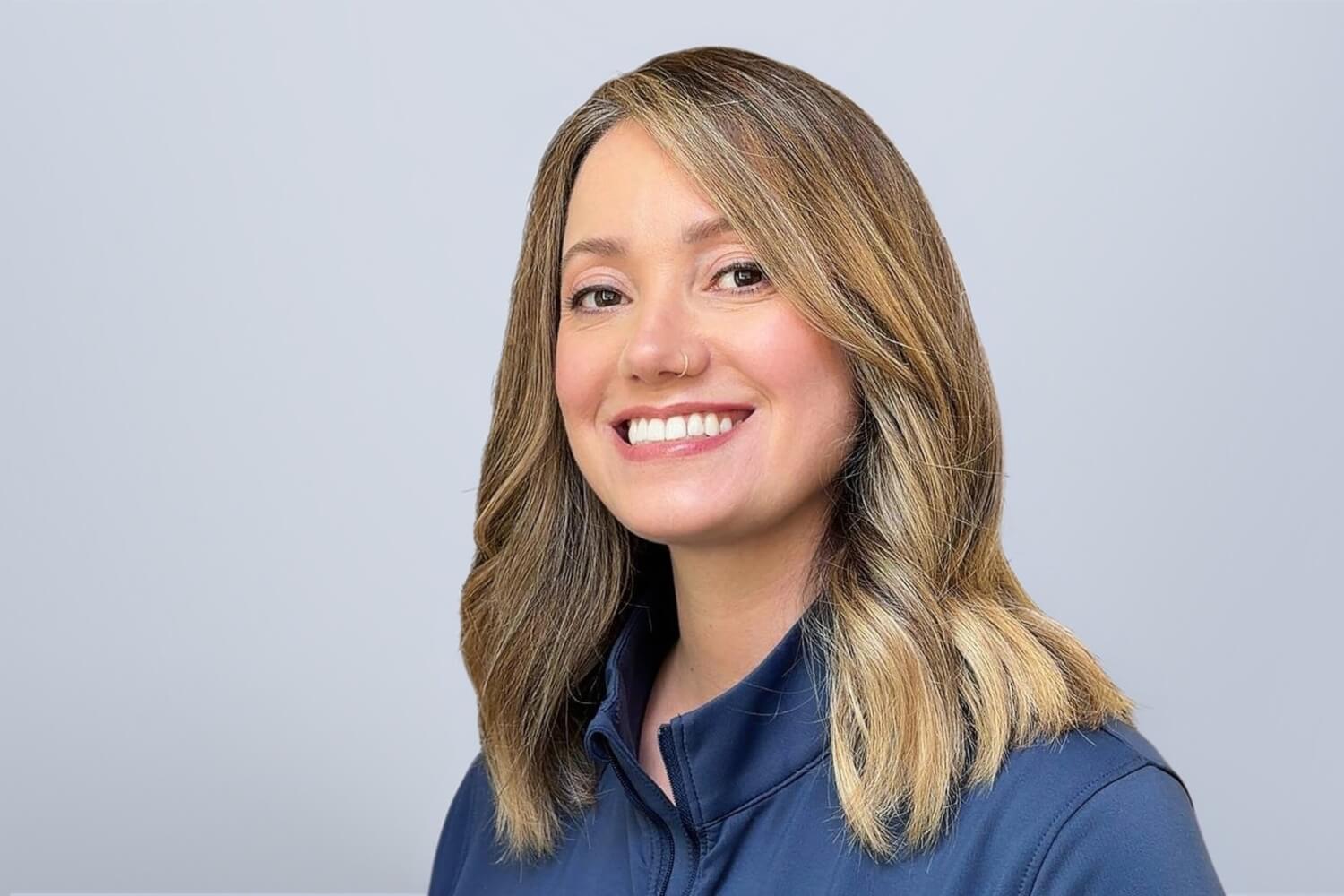 Smiling woman with shoulder-length light brown hair, wearing a blue collared shirt, posed against a plain light gray background.